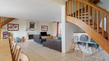 A spacious living room with a fireplace, a dining area with chairs, a staircase with wooden railings, and a baby play area with a changing station.