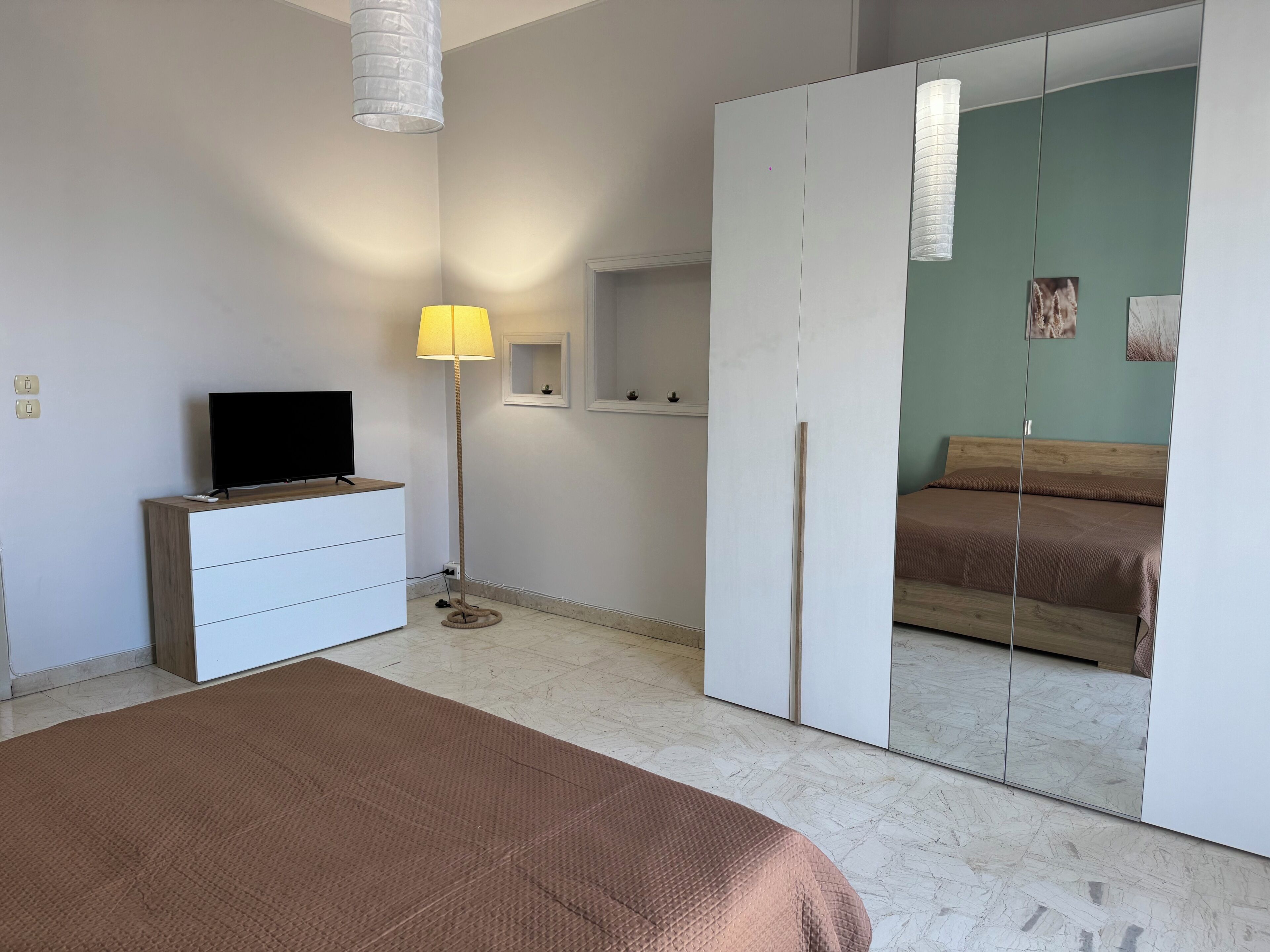A bedroom with a bed, a TV on a dresser, a floor lamp, and a wardrobe with a mirror.