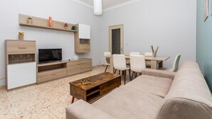 A modern living room with a flat-screen TV, a wooden coffee table, a beige sofa, and a dining area with chairs and a table. - Nicola House (Catania)