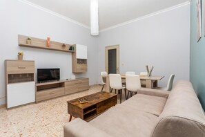 Ein modernes Wohnzimmer mit Flachbildfernseher, Couchtisch aus Holz, beiger Couch und Essbereich mit Stühlen und Tisch.