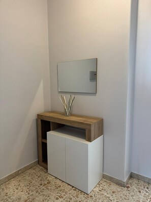 A corner room with a white cabinet, a wooden shelf with a vase, and a wall-mounted mirror.