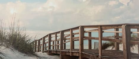 A wooden boardwalk leading to a beach with sand dunes and sparse vegetation.