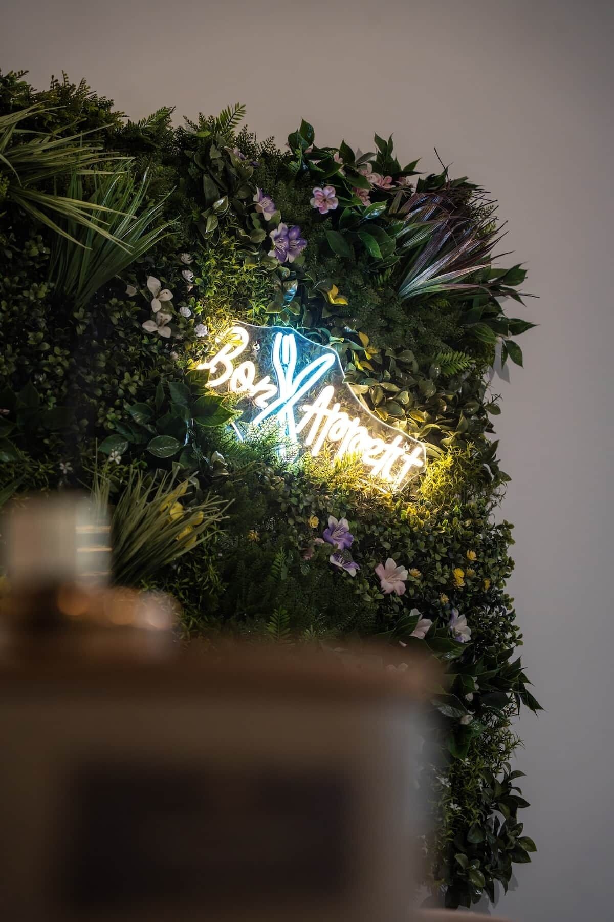 A neon sign with the text 'Be Xpert' surrounded by a lush green foliage arrangement.