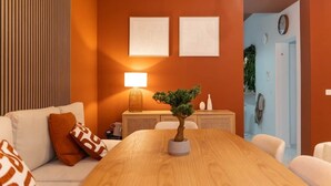 A modern living room with a wooden dining table, a sofa with patterned cushions, a lamp, and two framed artworks on the wall.
