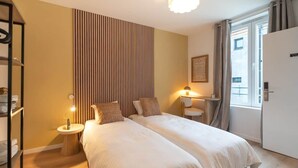 Habitación de hotel con dos camas, una mesita de noche con lámpara, un armario y una ventana con cortinas.