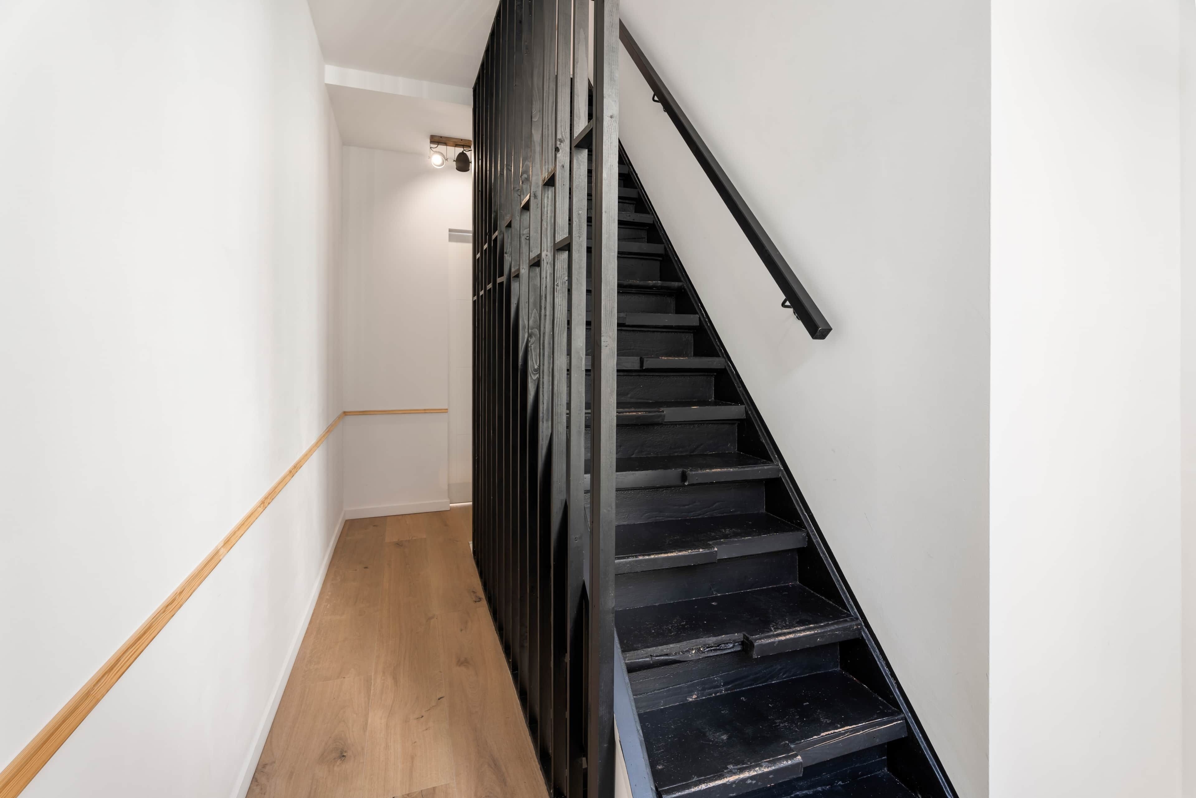 Un escalier avec des marches noires et une rampe en bois, menant à un palier où se trouve un luminaire.