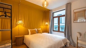 A hotel room with a bed, two bedside lamps, a window with curtains, a chair, and a small table. - Chez Thérèse (Bruxelles)