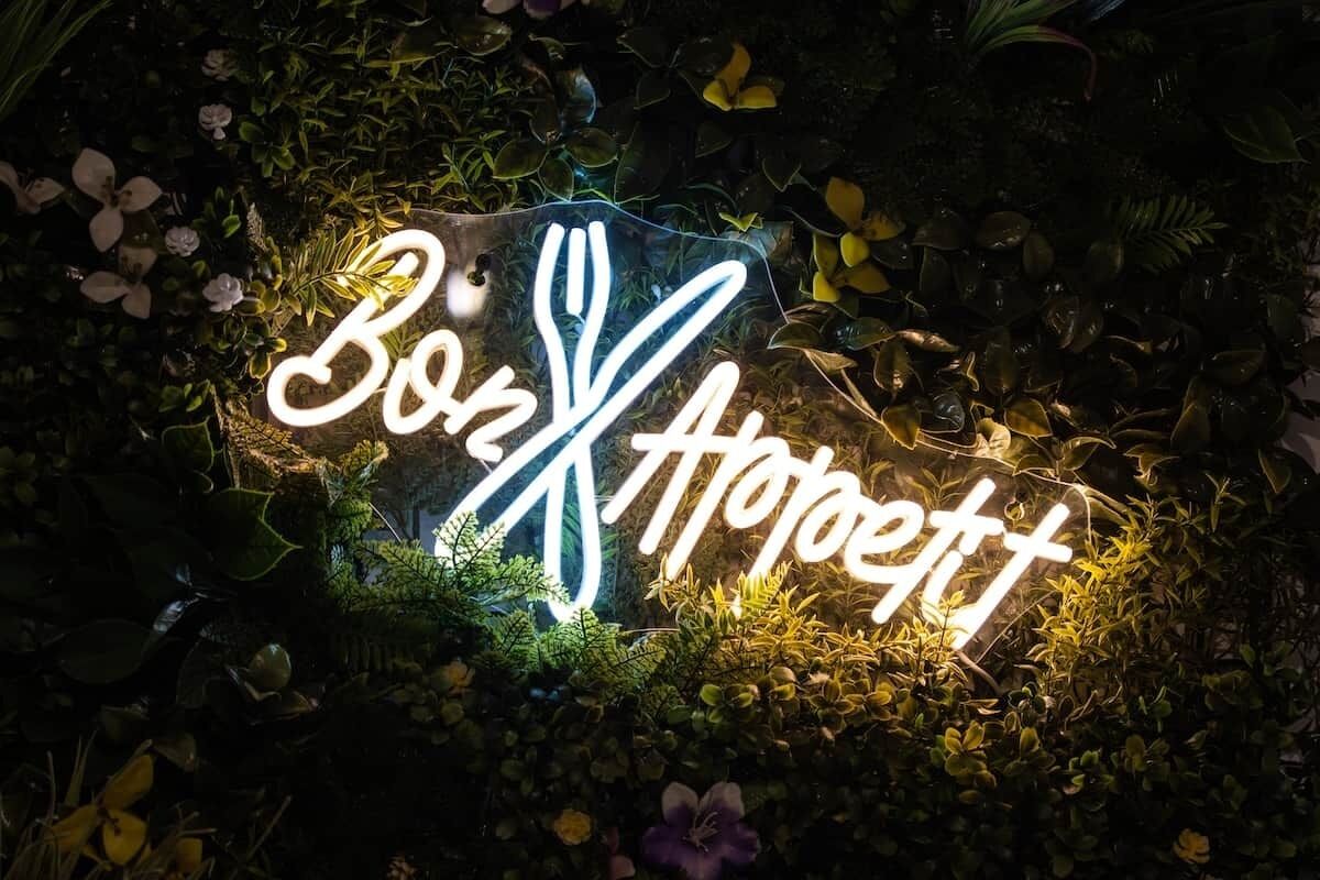 Een neonbord met de tekst 'Bon Appetit' omringd door begroeiing.