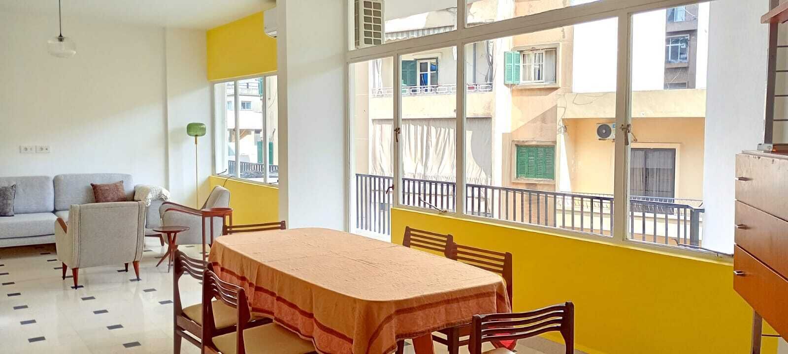 A room with a dining table, chairs, a sofa, a lamp, and a window with a balcony view.