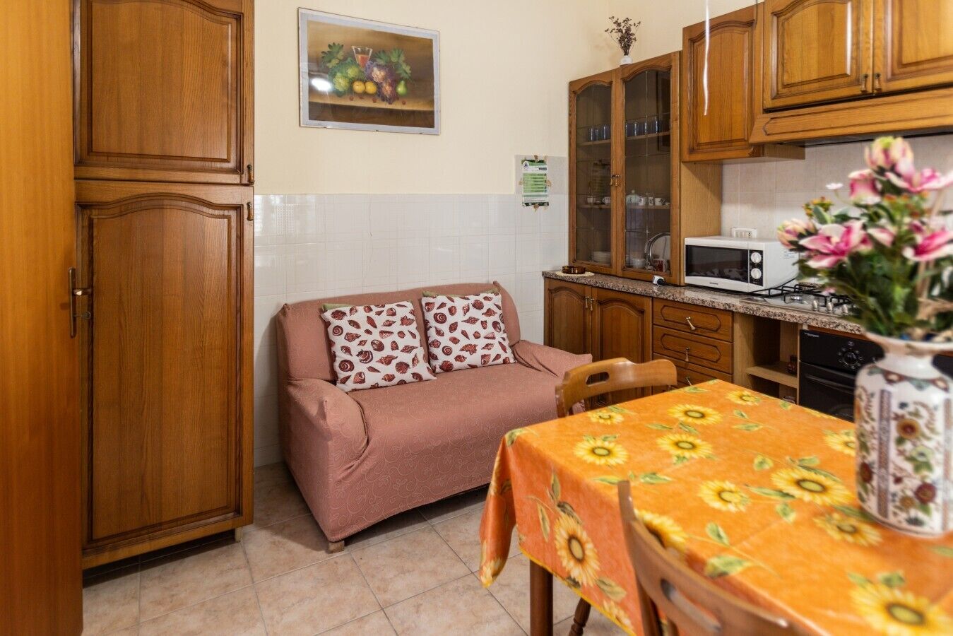 Una cucina compatta con mobili in legno, un forno a microonde, un tavolo da pranzo con tovaglia a fiori e un divano con cuscini decorati.