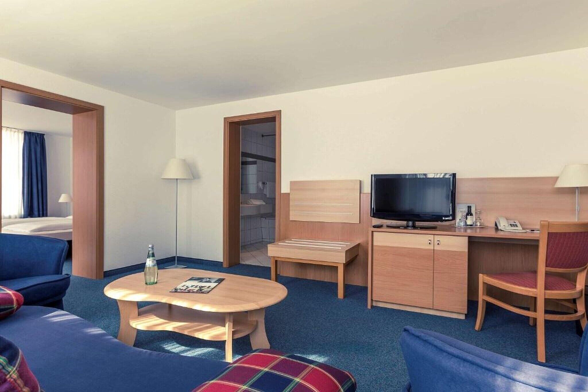 Et hotelværelse med en seng, et skrivebord med fjernsyn, en stol, et bord med en flaske og en lampe.