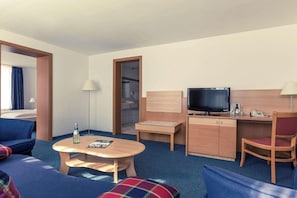 A hotel room with a bed, a desk with a TV, a chair, a table with a bottle, and a lamp.