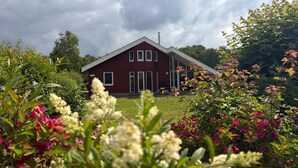En röd byggnad med ett sadeltak, omgiven av frodig grönska och blommande växter.