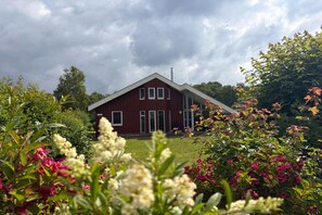 Ein rotes Gebäude mit Satteldach, umgeben von üppigem Grün und blühenden Pflanzen.