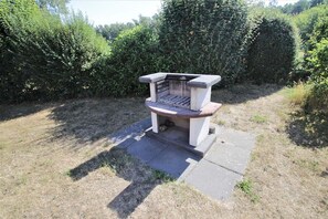 Eine Bank mit geschwungener Rückenlehne und Armlehnen, auf einer Betonplatte in einer grünen Umgebung mit dichter Strauchbepflanzung im Hintergrund.