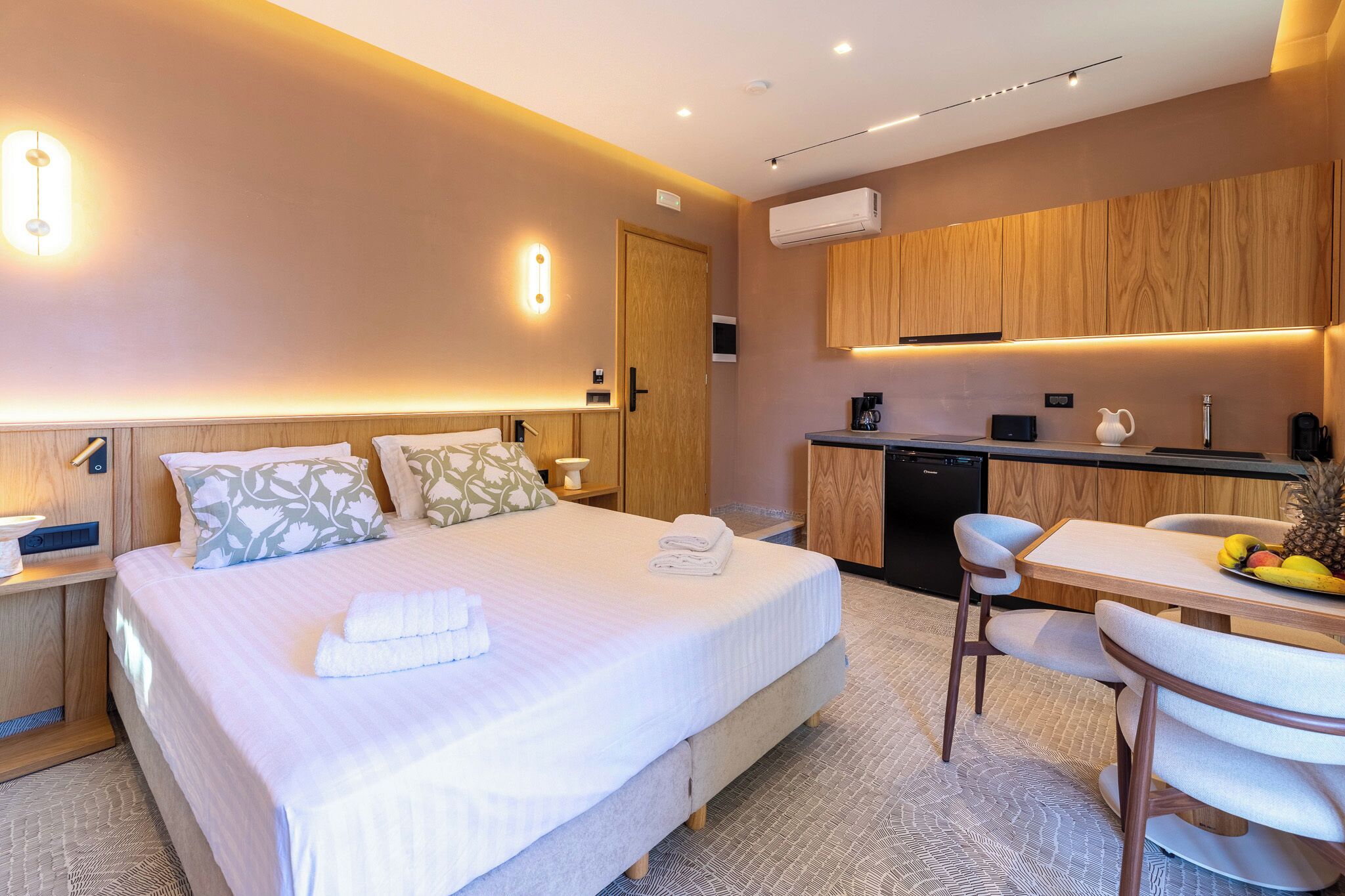 Een hotelkamer met een bed, nachtkastjes, een bureau met stoelen, een keukenhoek en een airconditioning.