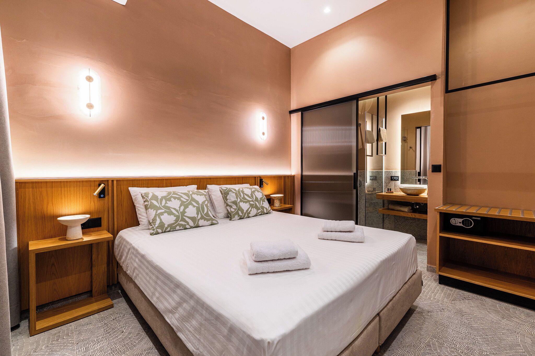 A modern hotel room with a large bed, wooden nightstands, a built-in TV, and a bathroom with a mirror and sink.