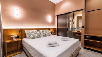 A modern hotel room with a large bed, wooden nightstands, a built-in TV, and a bathroom with a mirror and sink.