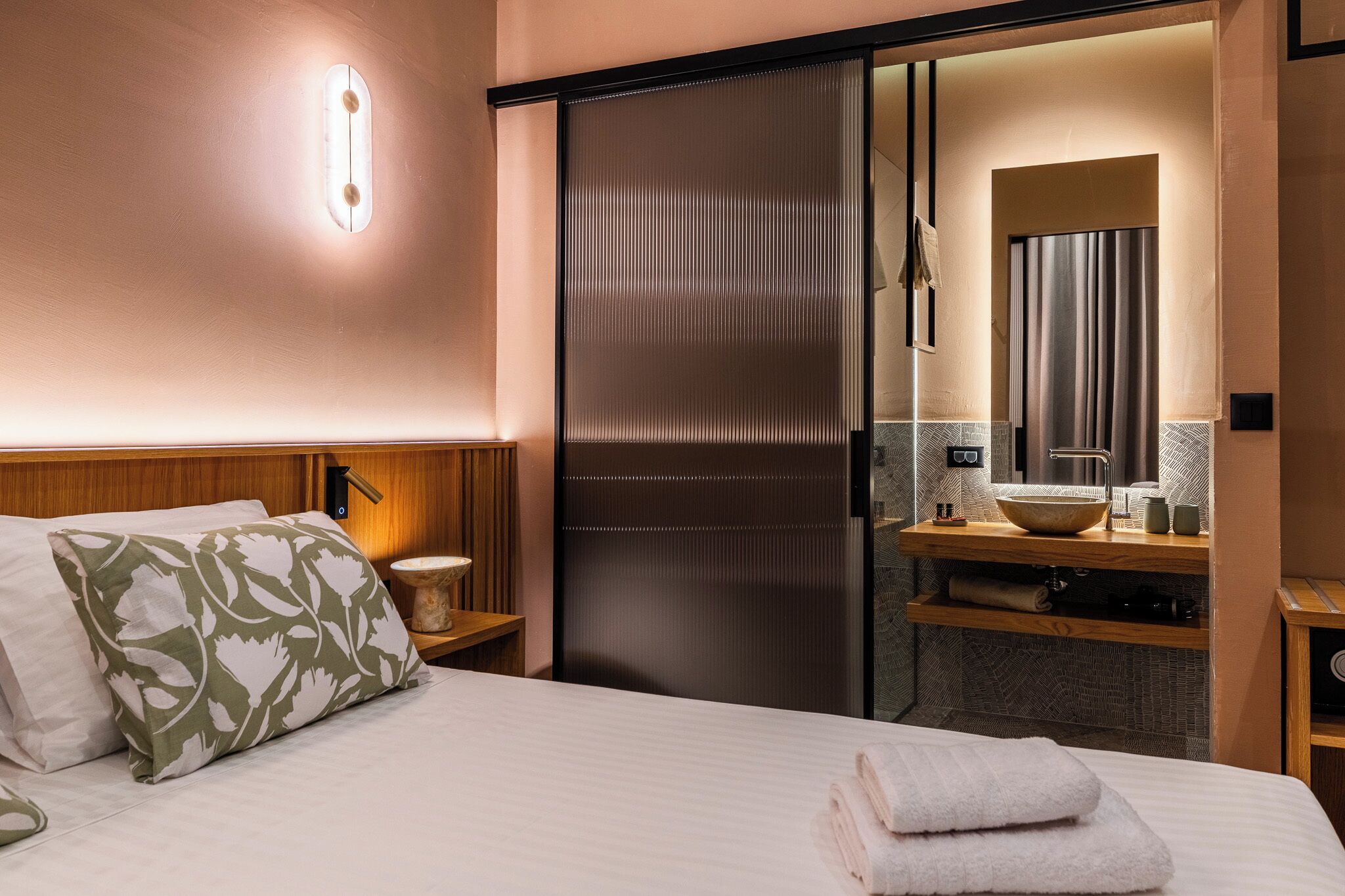 Een hotelkamer met een bed, een houten nachtkastje en een badkamer met een wastafel, spiegel en handdoekrek.