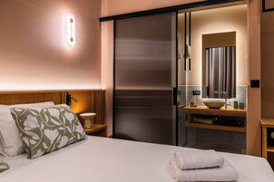 Habitación de hotel con cama, mesita de noche de madera, baño con lavamanos, espejo y toallero.