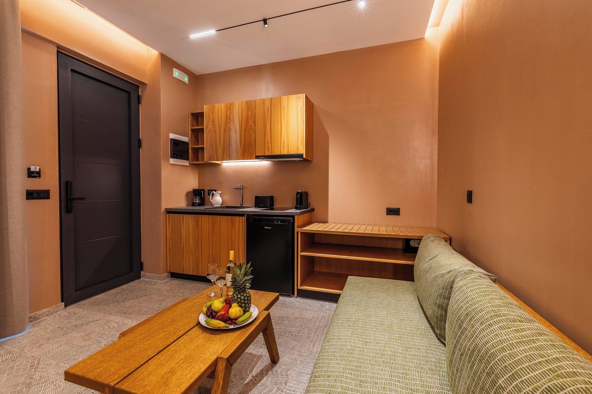 A compact living space with a kitchenette, a dining table with fruit, a sofa, and a door.