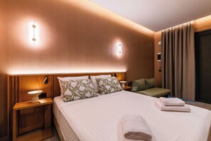 A hotel room with a bed, bedside tables, a nightstand, a wall-mounted light, a window with curtains, and a door. - Aetherion Suites (Chania)
