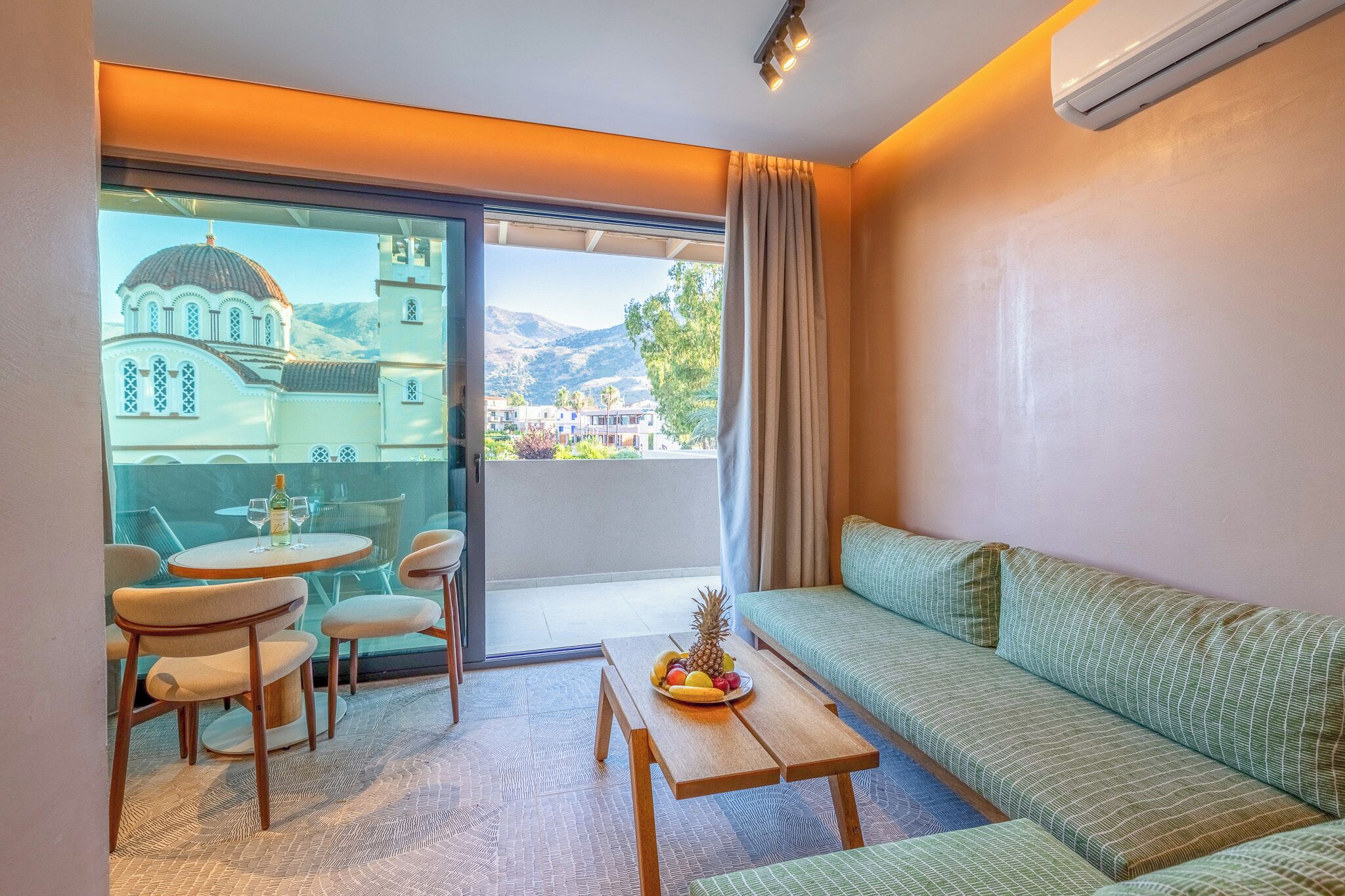 Una habitación moderna con un sofá, una mesa de comedor con sillas y un balcón con vistas a un edificio y montañas.