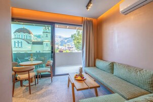 Una habitación moderna con un sofá, una mesa de comedor con sillas y un balcón con vistas a un edificio y montañas.