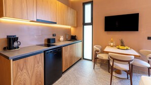 A modern kitchen with wooden cabinets, a dishwasher, a coffee maker, and a dining area with a table and chairs. - Aetherion Suites (Chania)
