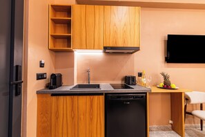 A compact kitchen with wooden cabinets, a built-in dishwasher, a coffee machine, and a small dining area with a chair and a fruit bowl. - Aetherion Suites (Chania)