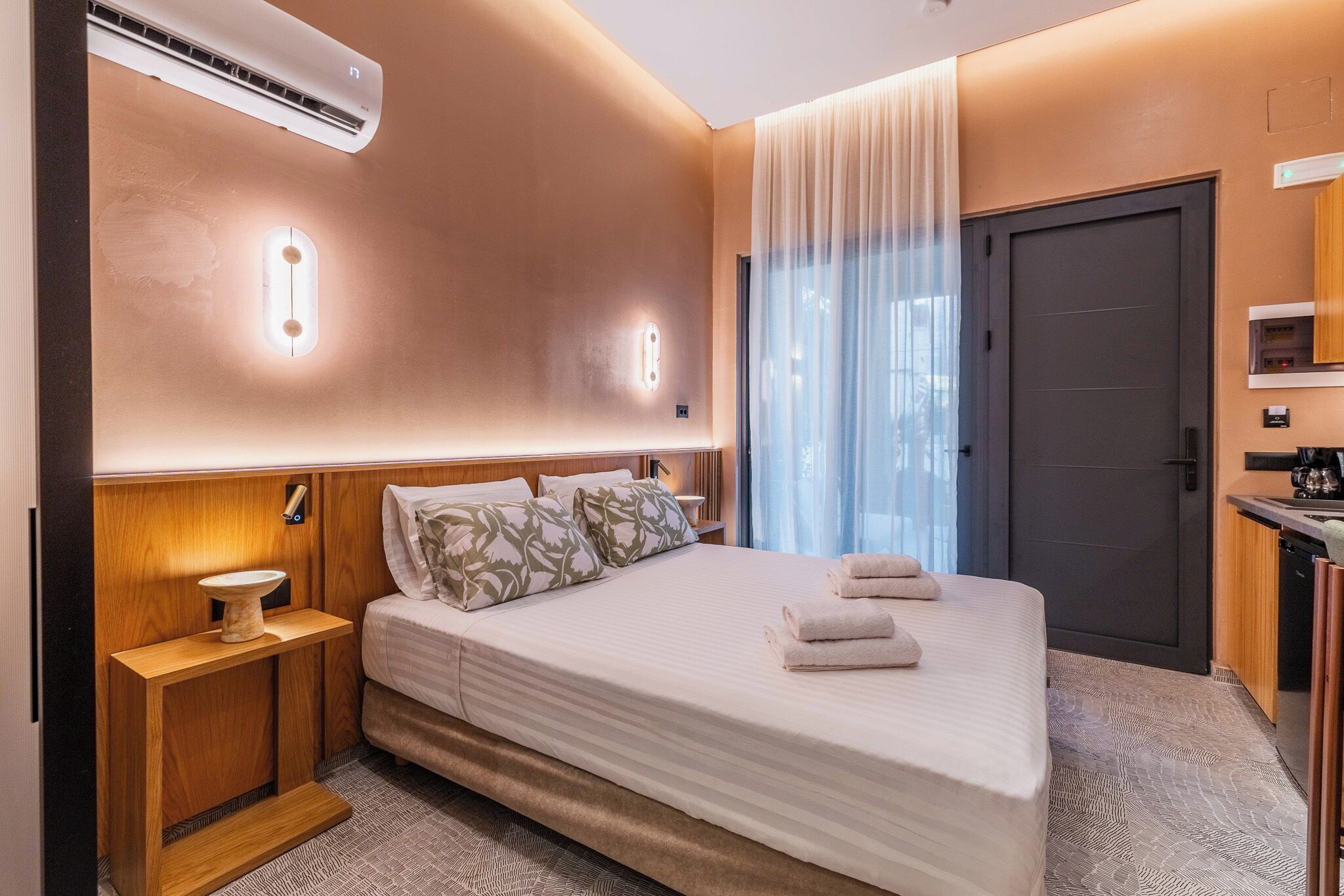 Ein modernes Hotelzimmer mit einem großen Bett, Nachttischen, einer Klimaanlage und Blick durch ein Fenster mit leichtem Vorhang.