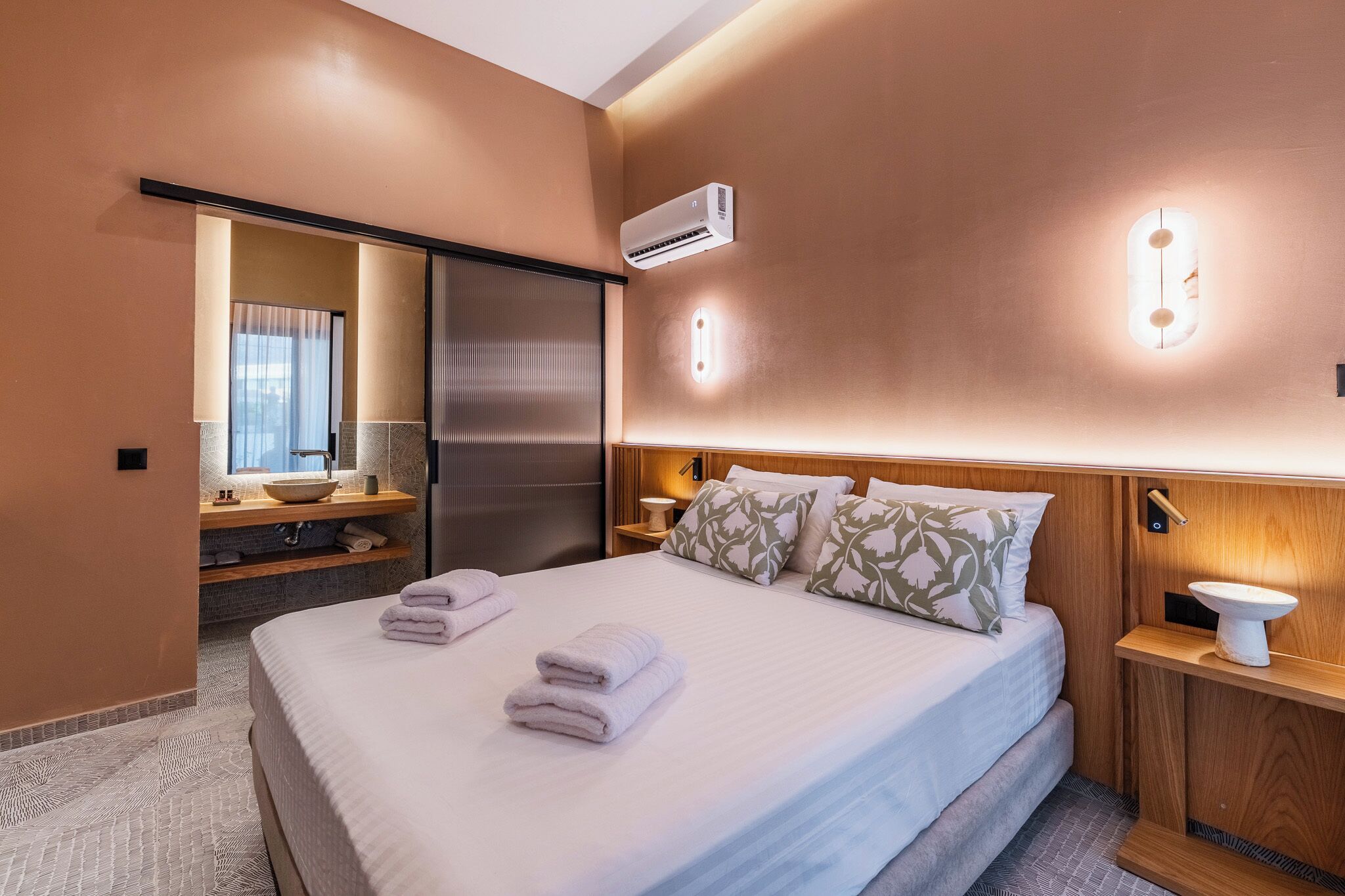 Habitación de hotel con cama, mesita de noche, espejo, ventana y aire acondicionado.