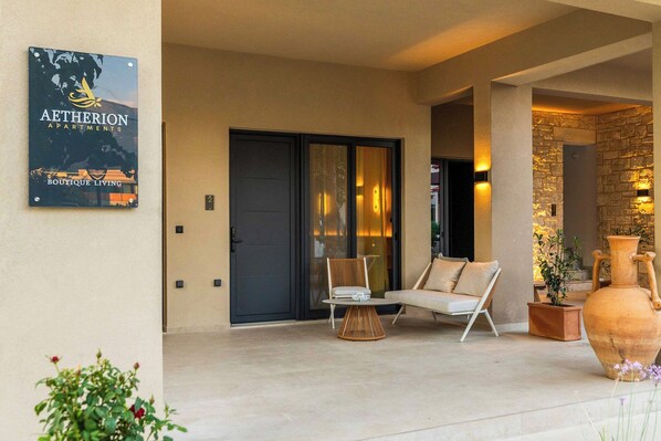 A modern outdoor seating area with a sofa and chairs, a large vase, and a sign for Aetherion Apartments. - Aetherion Suites (Chania)
