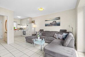 A modern living room with a grey sectional sofa, a glass coffee table, and a kitchen in the background. - Enjoy a relaxing getaway at the "Küstenzeit SPO" vacation apartment. (Sankt Peter-Ording)