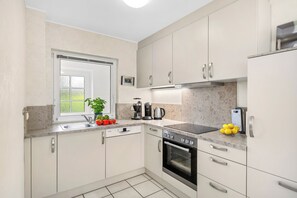 A modern kitchen with white cabinets, a built-in oven, and a window with a view of greenery. - Enjoy a relaxing getaway at the "Küstenzeit SPO" vacation apartment. (Sankt Peter-Ording)