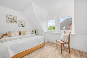 A bedroom with a sloped ceiling, a bed with a white quilt, two windows, and a wooden chair. - Enjoy a relaxing getaway at the "Küstenzeit SPO" vacation apartment. (Sankt Peter-Ording)
