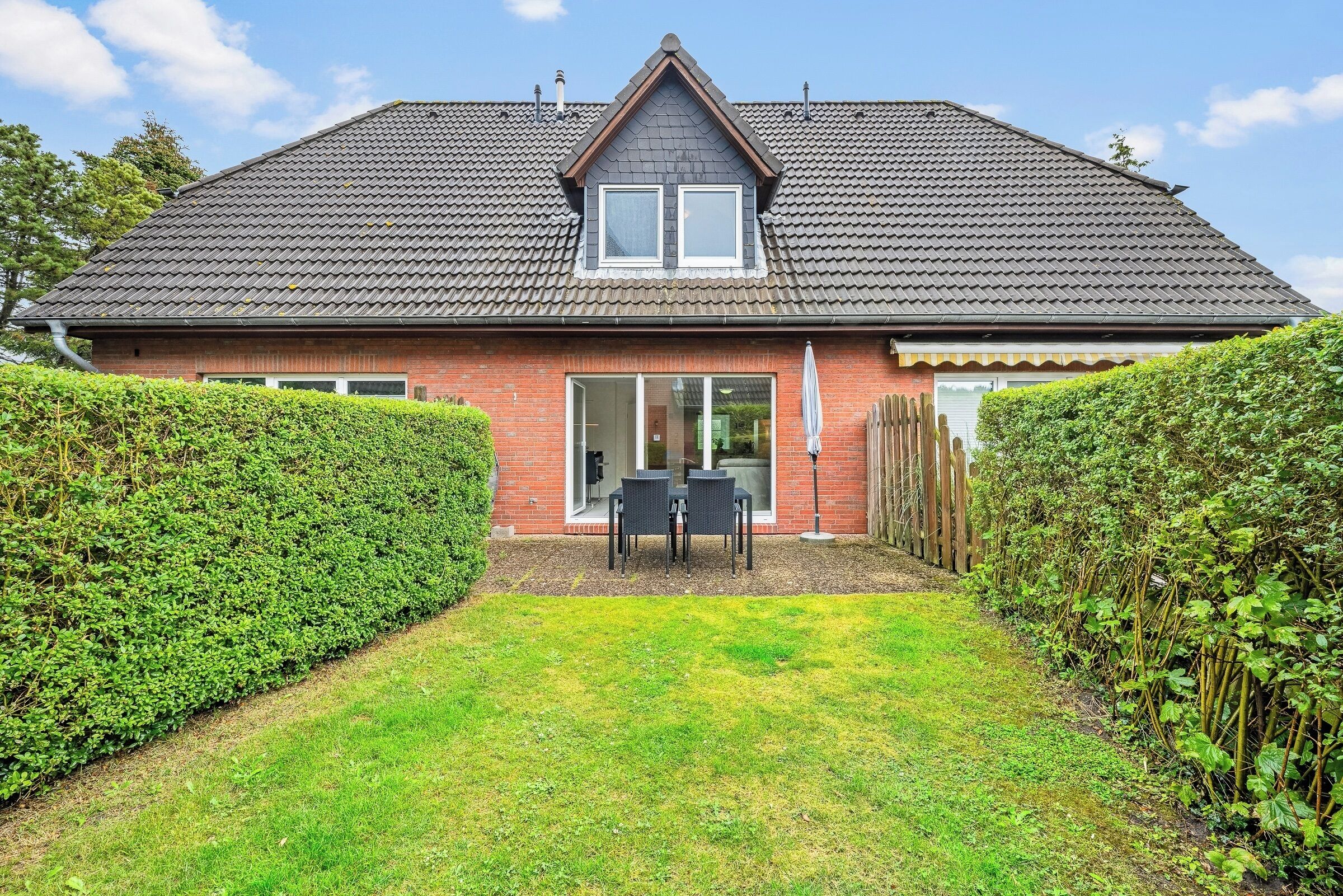 Une maison à deux étages, avec un toit en tuiles, une porte d’entrée et un petit patio aménagé avec du mobilier d’extérieur.
