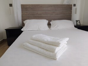 Un lit bien fait, avec du linge de lit et des serviettes blanches, une tête de lit en bois et deux tables de chevet, dont l’une est ornée d’un tableau encadré.