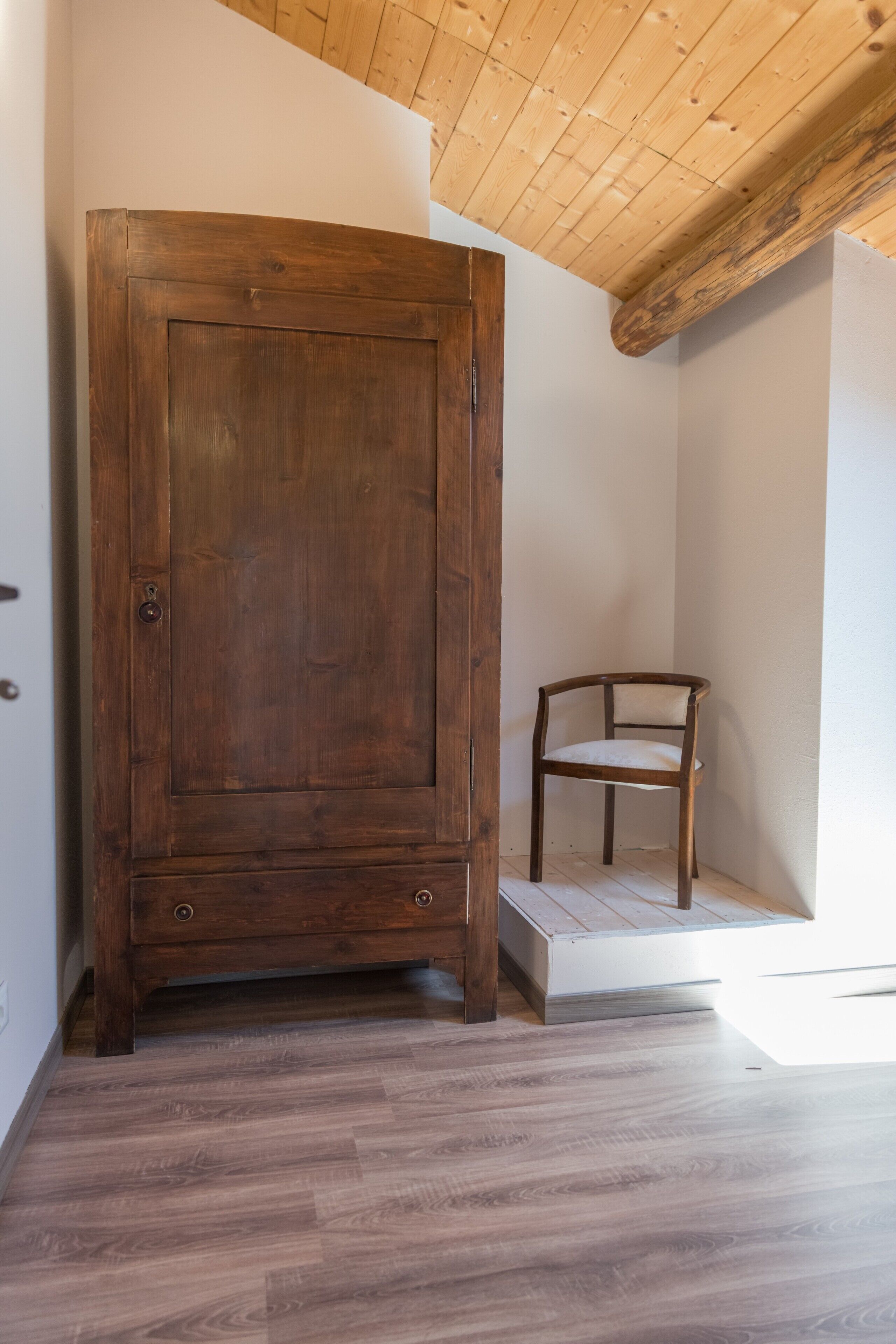 Un armario de madera con una pequeña plataforma elevada y una silla en una esquina de una habitación con suelo de madera y techo inclinado.