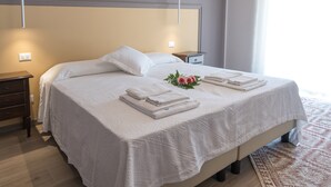 Een opgemaakt bed met witte lakens en een bloemstuk, een nachtkastje met een lamp, en een houten vloer.