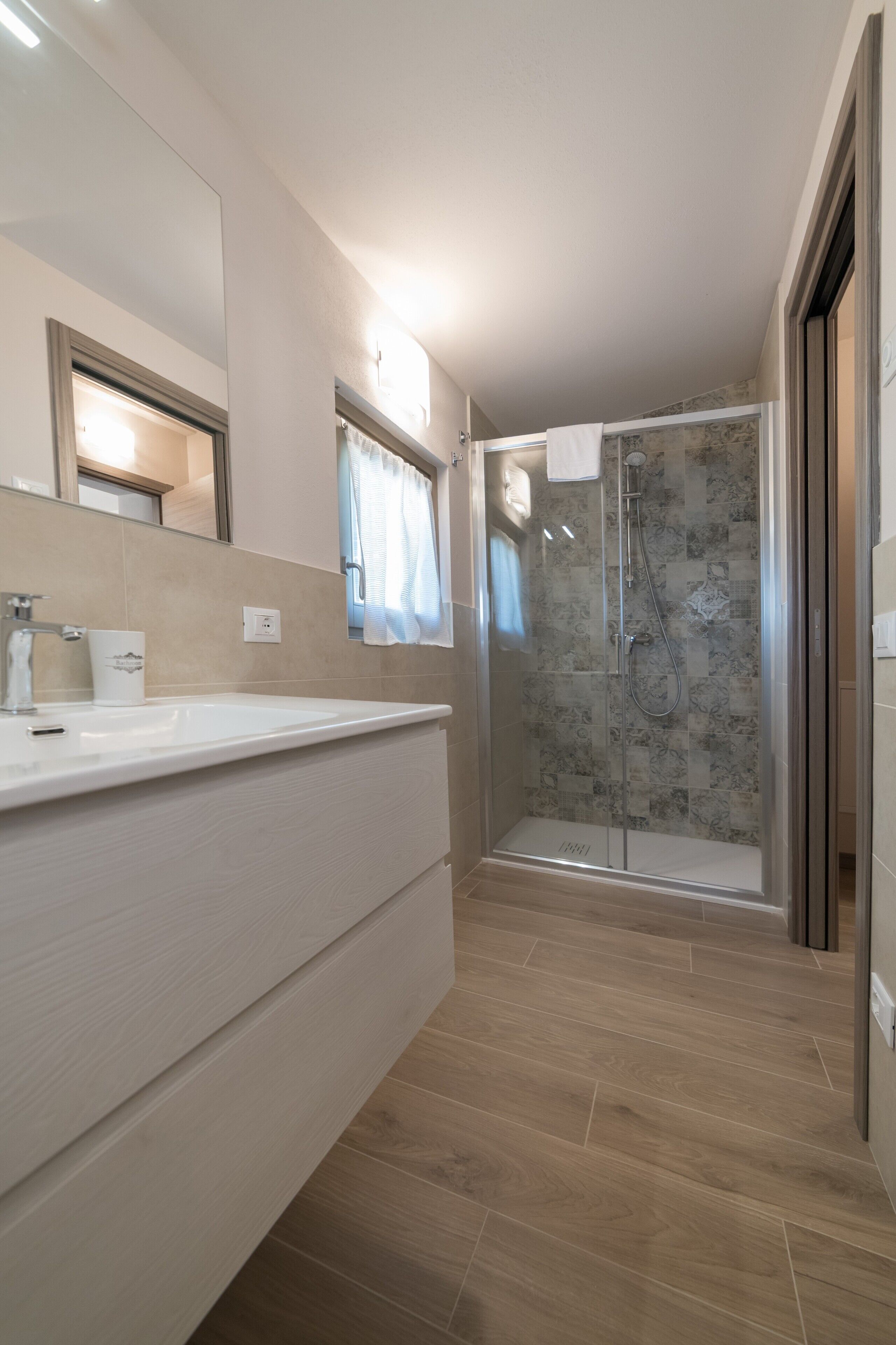 Un bagno moderno con uno specchio grande, un lavandino bianco, una doccia con porta in vetro e pavimento in legno.