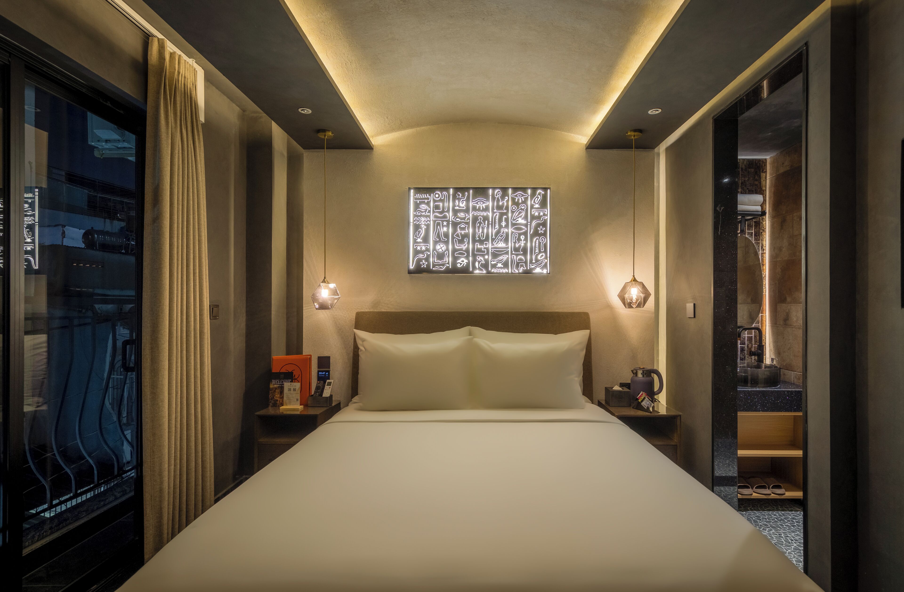 Quarto de hotel com uma cama grande, mesa de cabeceira com abajur, janela com cortinas e uma obra de arte na parede.