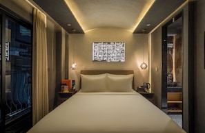Habitación de hotel con una cama grande, una mesita de noche con lámpara, una ventana con cortinas y una obra de arte en la pared.