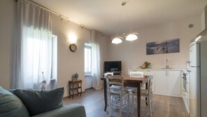 Una sala de estar moderna con sofá, mesa de comedor y sillas, una cocina con armarios y electrodomésticos blancos, y un cuadro en la pared.