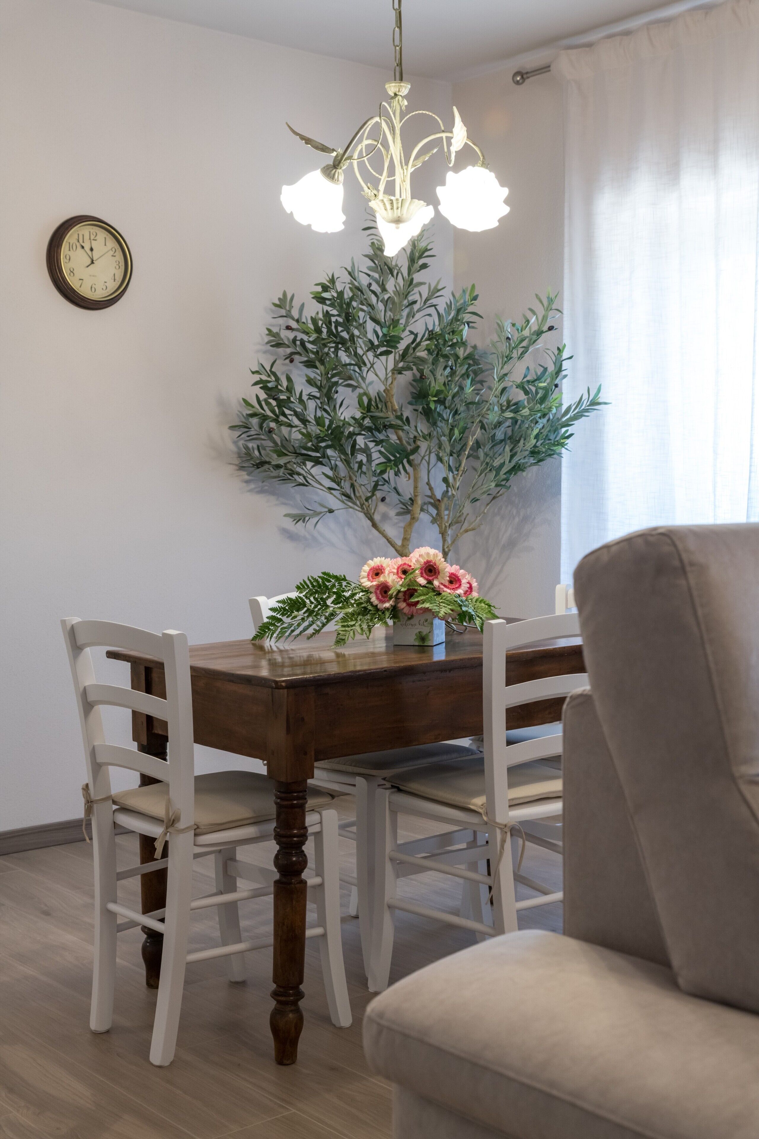 Ein Essbereich mit Holztisch, weißen Stühlen, einer Chandelier-Leuchte, einer Uhr und einem Blumentopf.