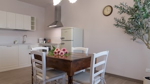 Uma área de jantar com uma mesa de madeira e cadeiras brancas, um lustre pendurado, um relógio na parede e um vaso com planta.