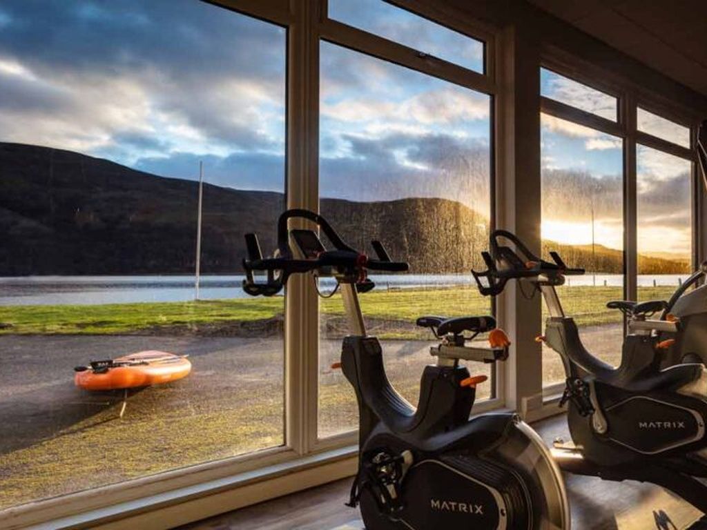 Ein Indoor-Cycling-Bereich mit zwei Ergometer, einem Rudergerät und Blick auf einen See und Berge durch große Fenster.