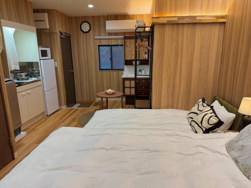 A compact hotel room with a bed, bedside table, and a kitchenette area.