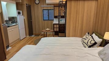 A compact hotel room with a bed, bedside table, and a kitchenette area.