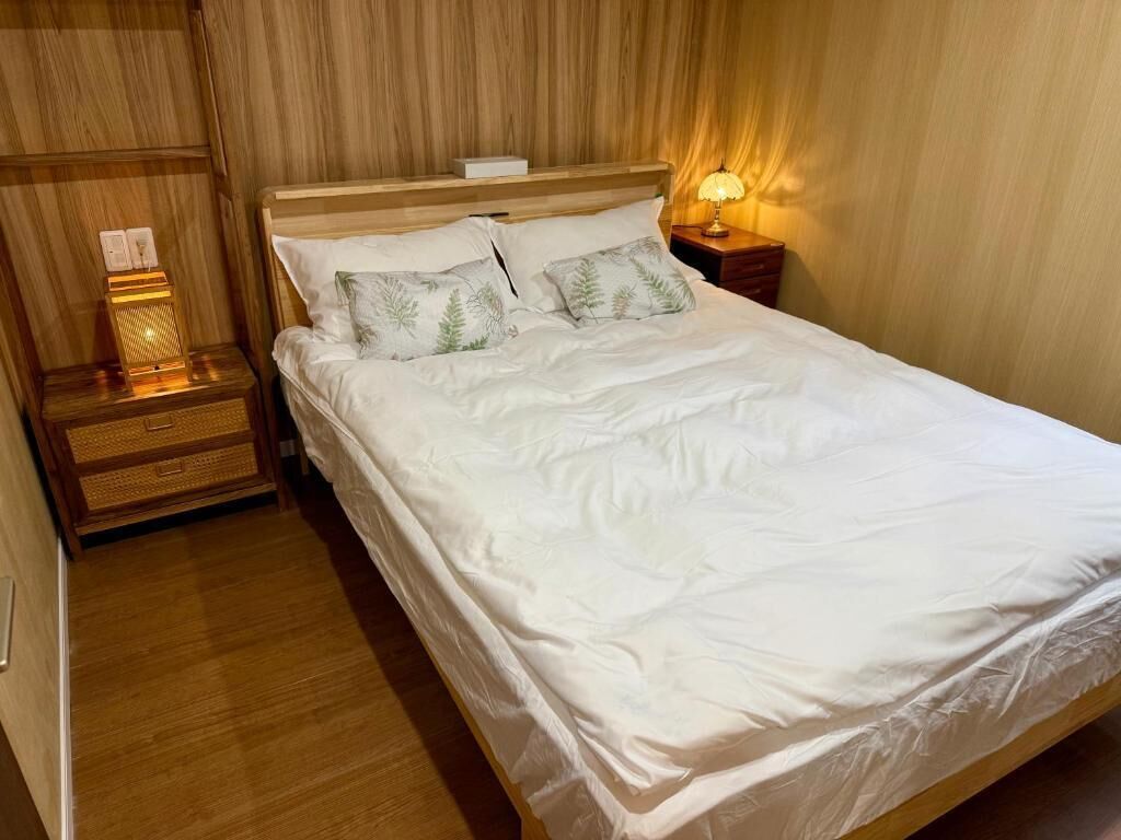 Una cama bien tendida con sábanas blancas y dos almohadas decorativas, una lámpara de noche y una mesita de madera con una cesta tejida.